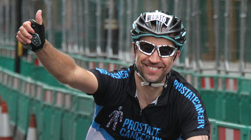 Man in cycling helmet, smiling with sunglasses on. His right arm is gesturing with a thumbs up sign. He is wearing a black cycling jersey with the prostate cancer logos in bright blue. 