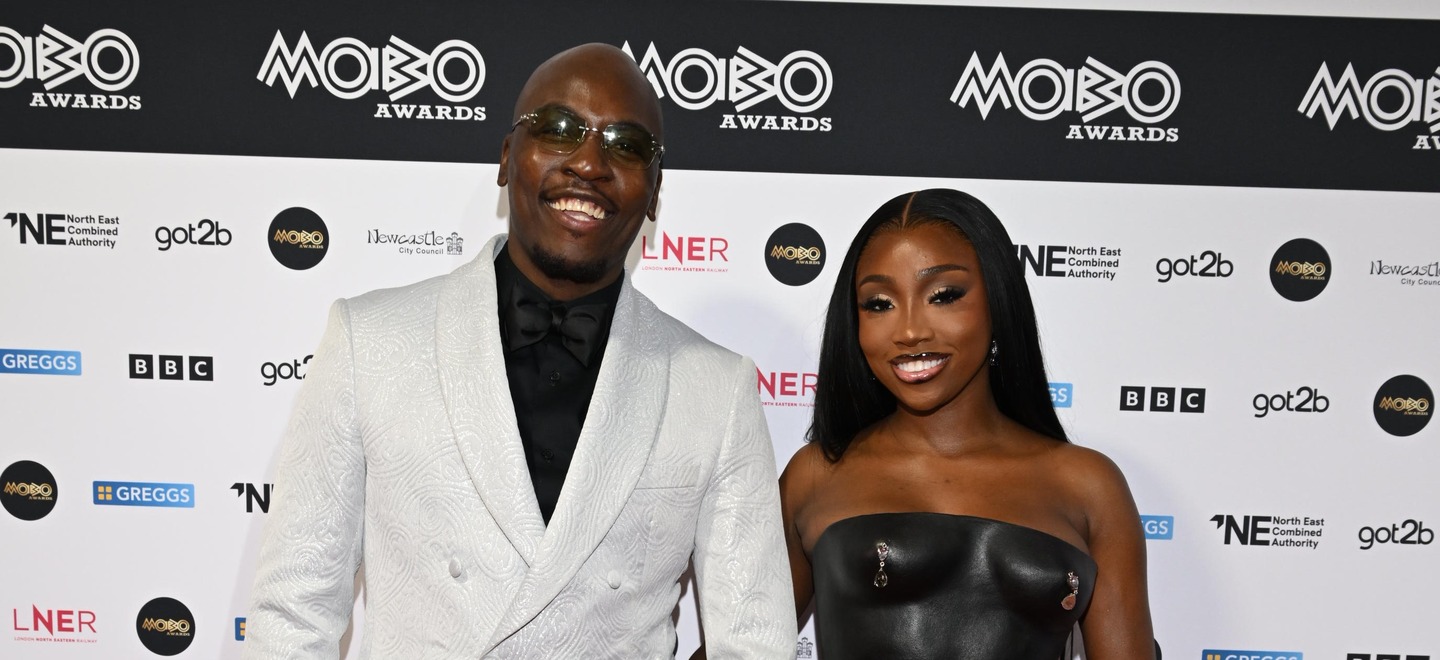 A man wearing sunglasses and a white suit jacket next to a woman wearing a black dress.