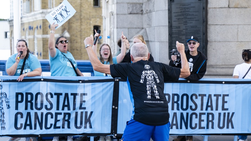 A man wearing a Prostate Cancer UK running top facing a row of supporters who are cheering for him next to a Prostate Cancer UK banner