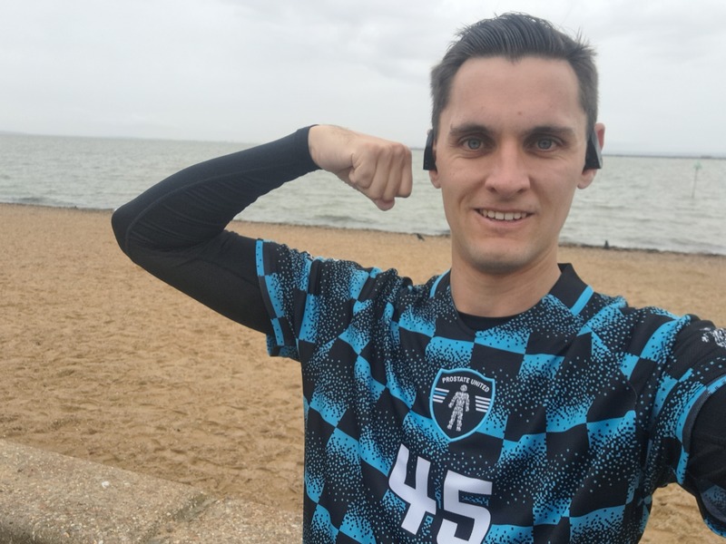 Charlie standing on a beach in his running kit, wearing a blue and black Prostate United shirt