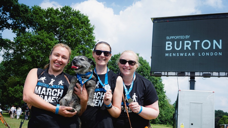 Three women and a dog smile in front of a promotional display for Burton Menswear.