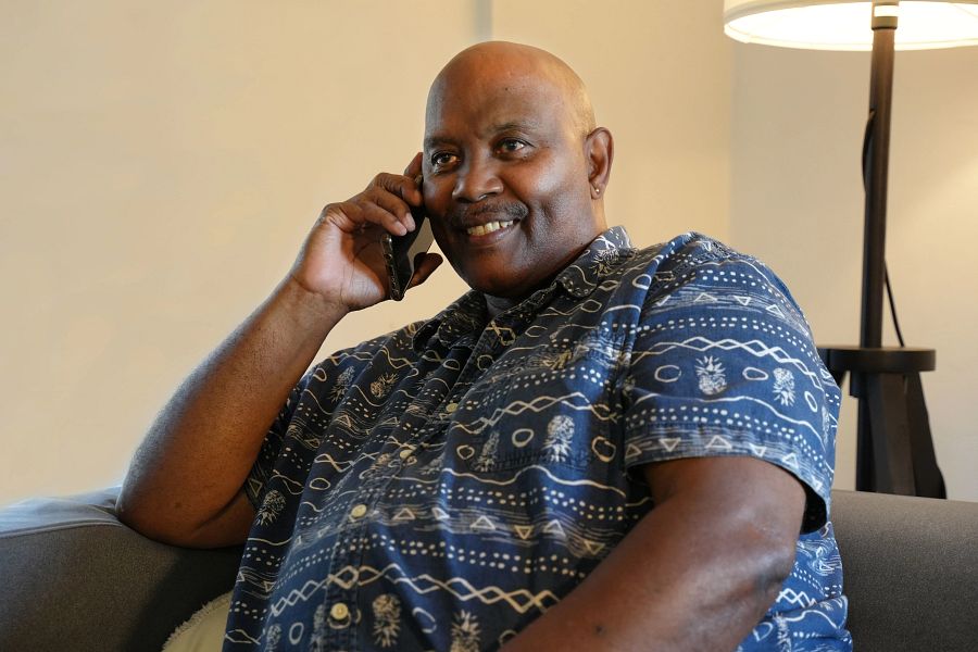 A man wearing a blue shirt smiling while sitting on a sofa and talking on the phone. 