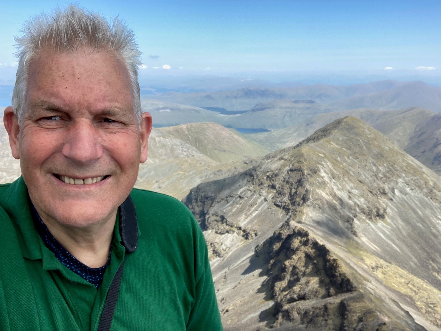 A man wearing a green top smiling at the camera in front of a mountainous background