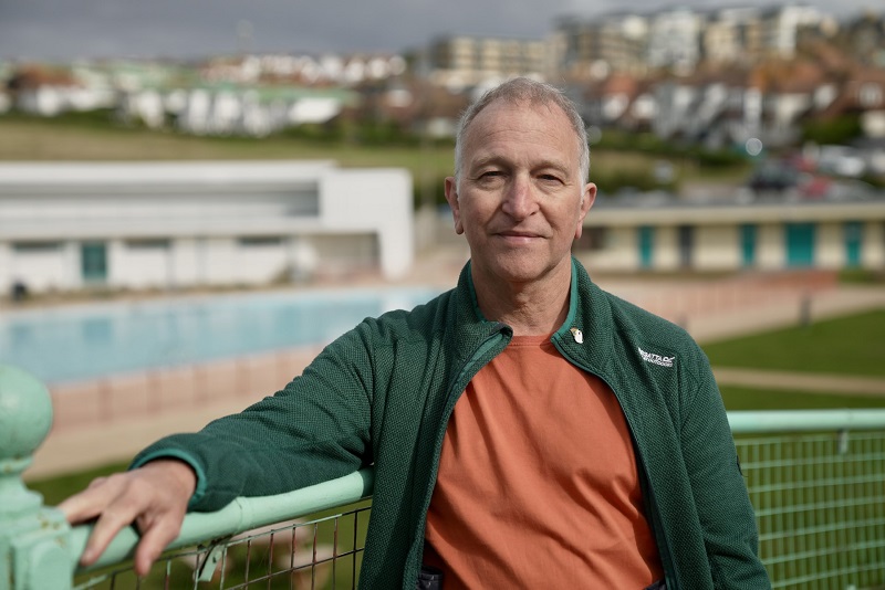 Giles, a man wearing an orange t-shirt and green jacket, stands in front of a blurry town background, looking at the camera.