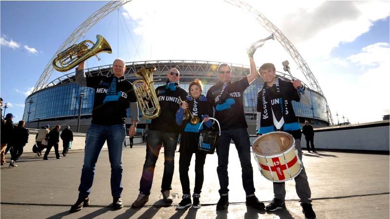 Five adults wearing Prostate United tee-shirts, standing in front of a football stadium and holding musical instruments