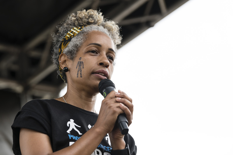 A woman speaking into a microphone on stage at a large public fundraising event in a park.