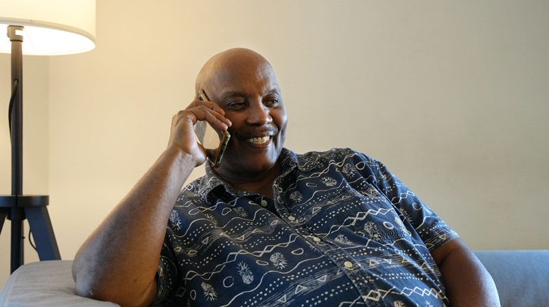 A man sitting on a sofa, talking on a phone and smiling