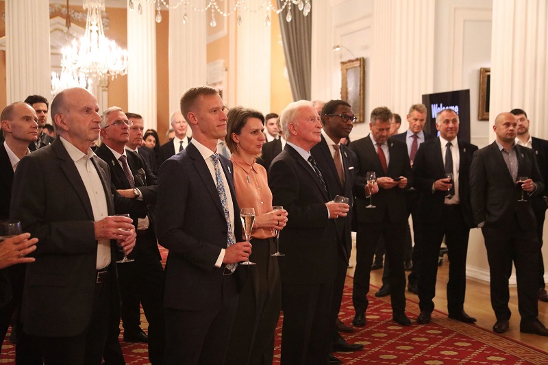 A group of people wearing suits and holding champagne flutes, looking towards a speaker
