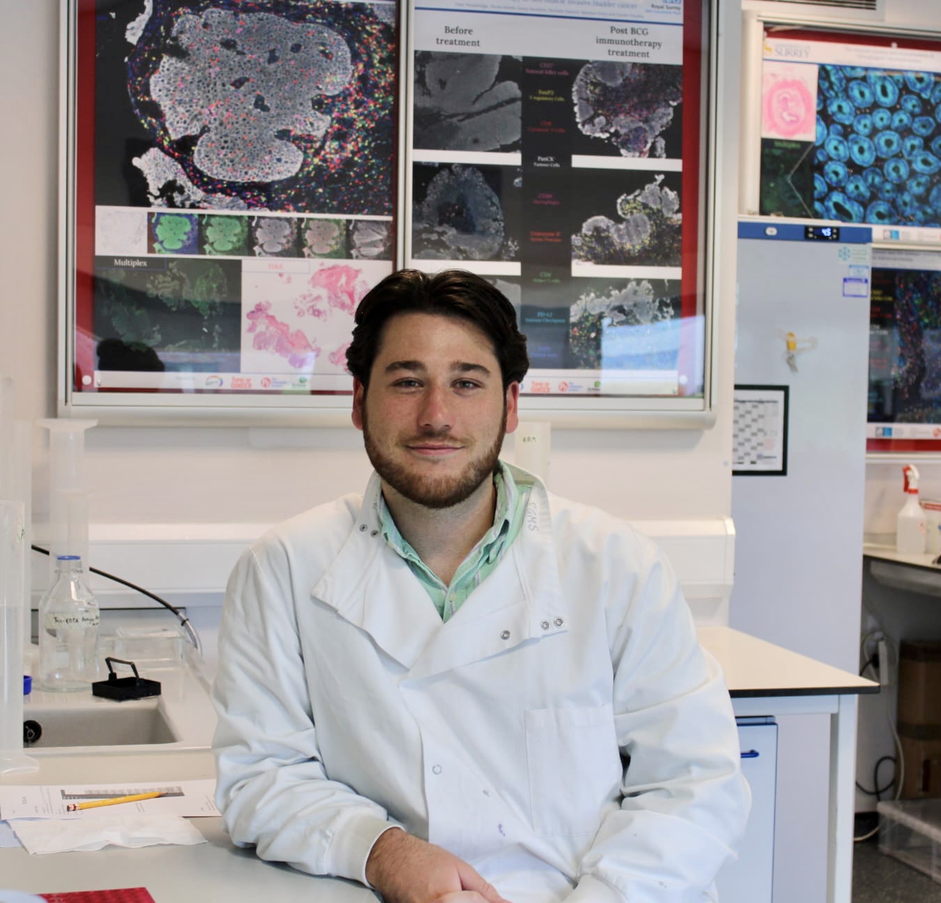A man with dark brown hair and a short beard wearing a white coat, standing in a lab