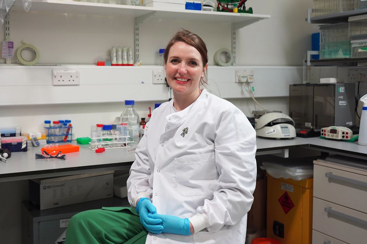 A woman with her hair tied back wearing a white lab coat and blue gloves, sitting in a lab and smiling