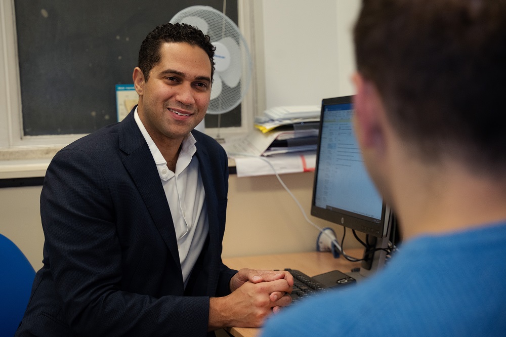 A man in a black suit and white shirt sitting by a computer, facing another person wearing blue clothing and smiling