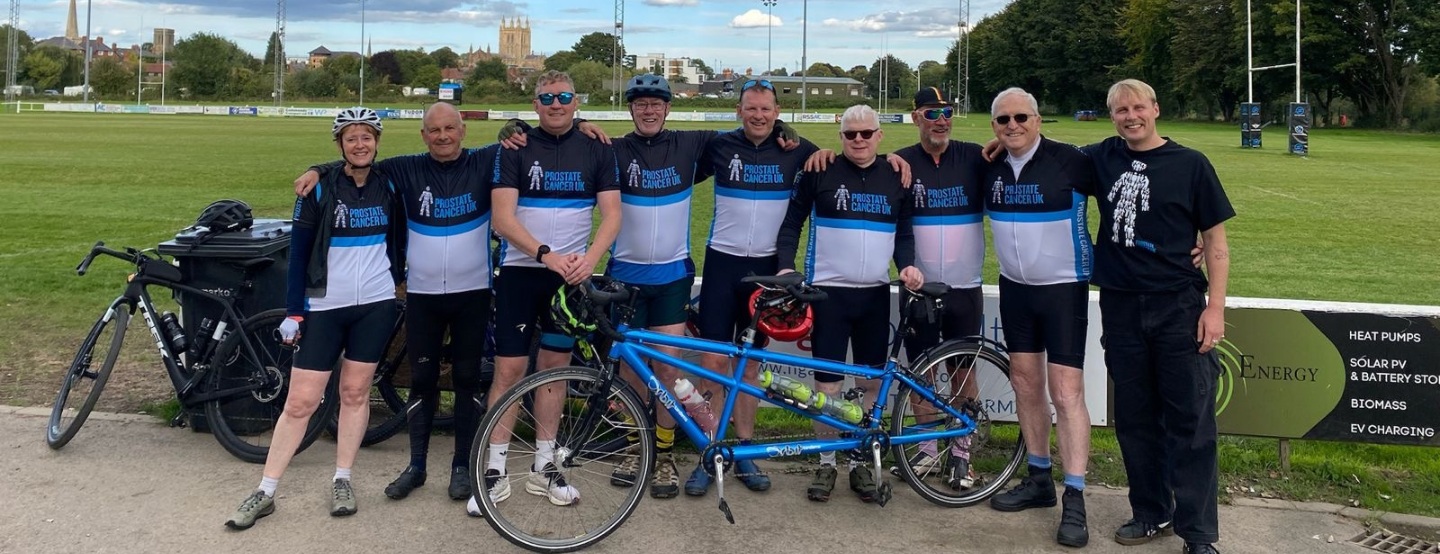 A group of cyclists wearing Prostate Cancer UK branded uniform, standing arm-in-arm in front of a playing field.