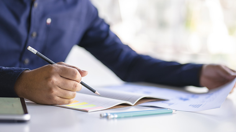 A man wearing a blue shirt writing with a pencil. 