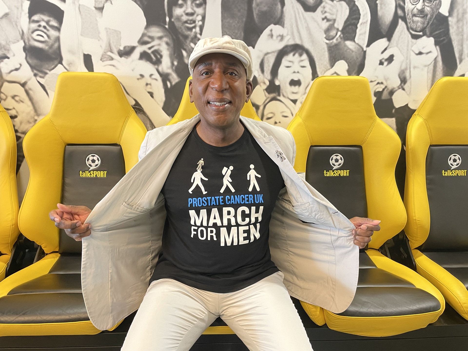 A man wearing a white suit jacket and white cap holding his jacket open to reveal a black Prostate Cancer UK t-shirt