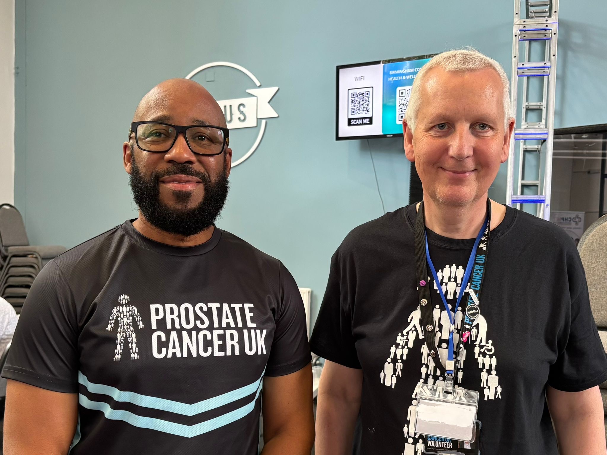 Two men wearing black Prostate Cancer UK t-shirts standing next to each other and smiling at the camera. 