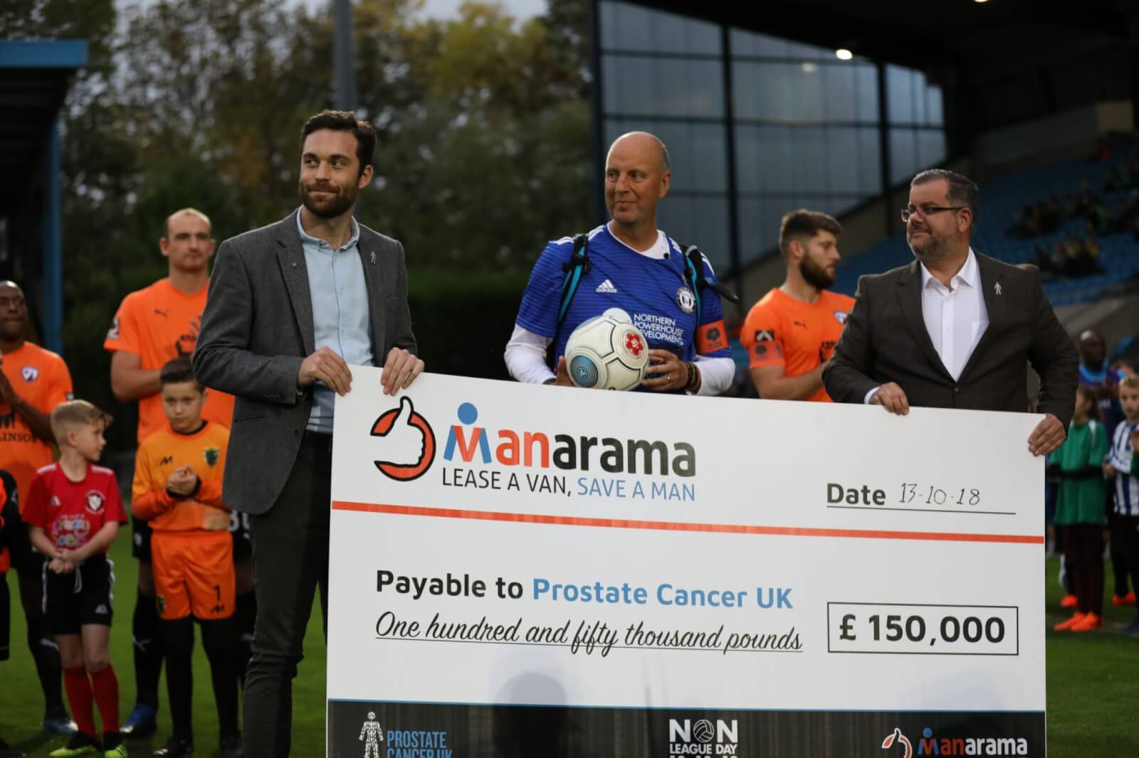 A man in a blue shirt stands next to two other men in suits holding up a giant check at the start of a football match.