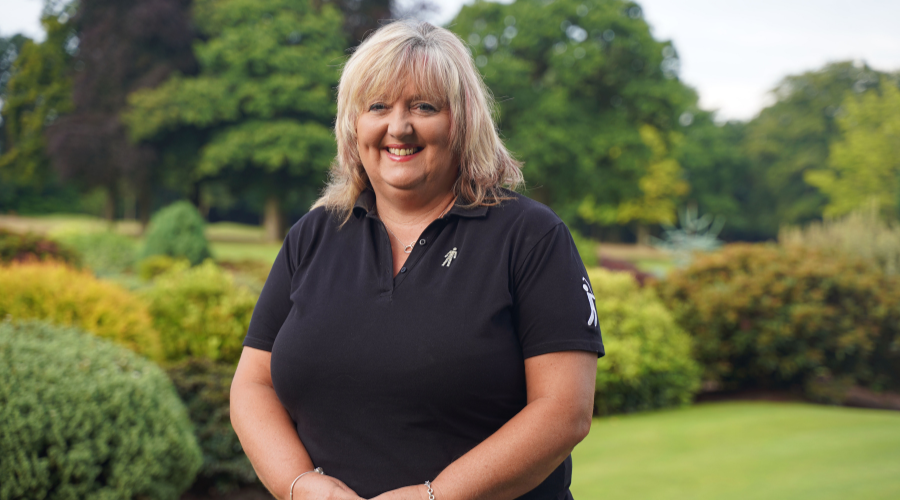 A woman with blonde hair standing on a golf course, wearing black golfing kit and a Prostate Cancer UK Man of Men badge.