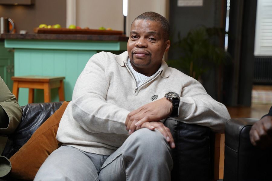 A Black man wearing a white sweater and grey trousers, sitting on a sofa with his hands resting on his knee