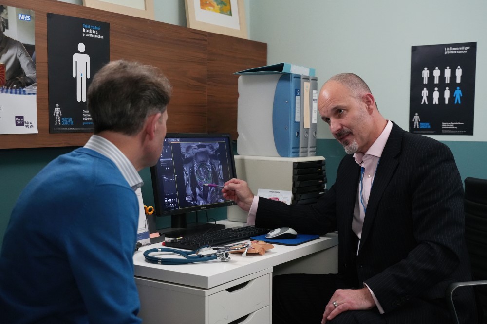 A male doctor wearing a black jacket gestures to a computer screen displaying a medical scan while talking to a man wearing a blue jumper.