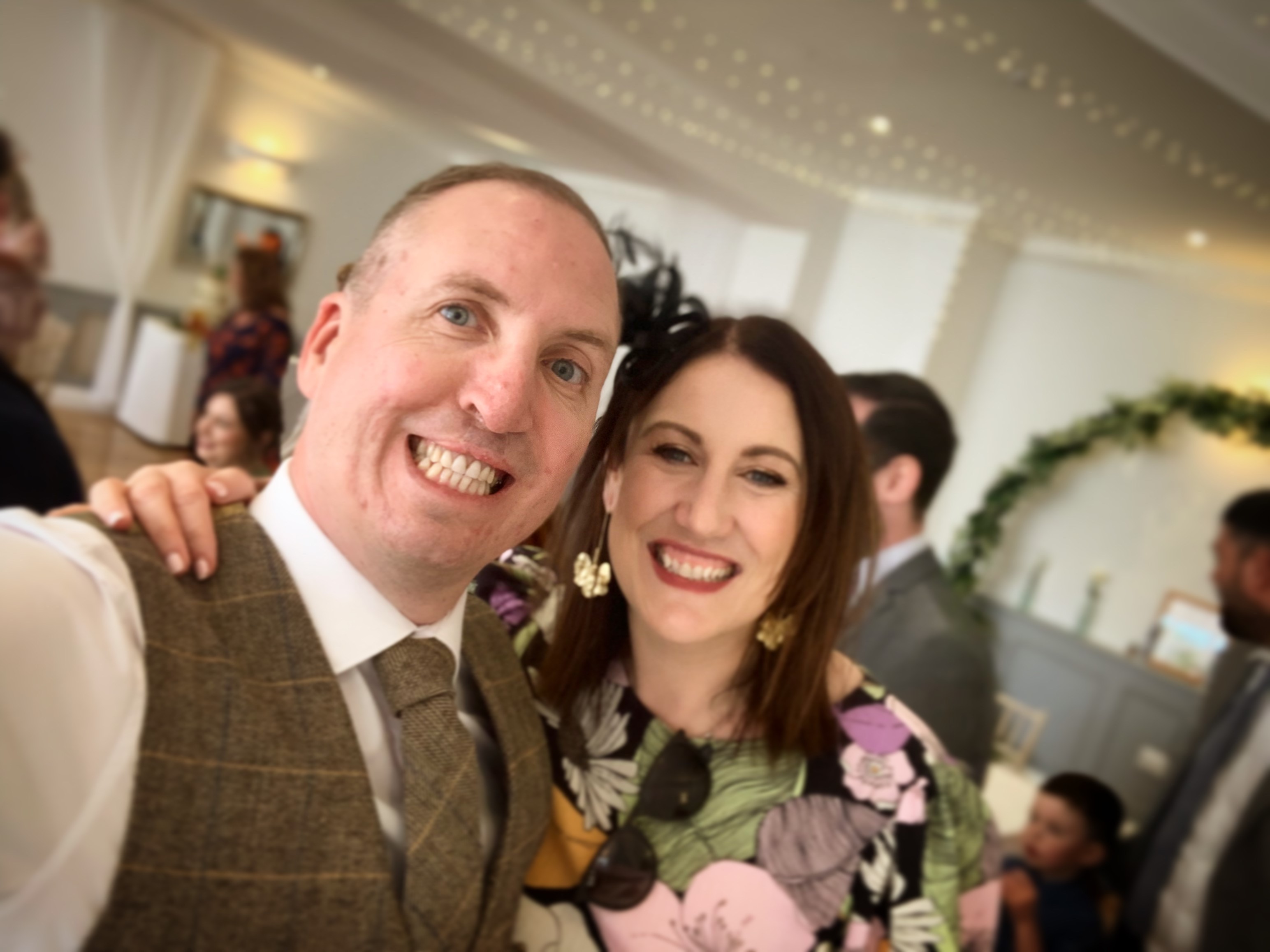 A smiling man and woman, dressed in smart clothes at a wedding, with their arms round each other's shoulders.