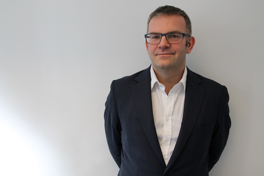 Professor Mark Emberton wearing glasses, a white shirt and black blazer, standing in front of a grey background