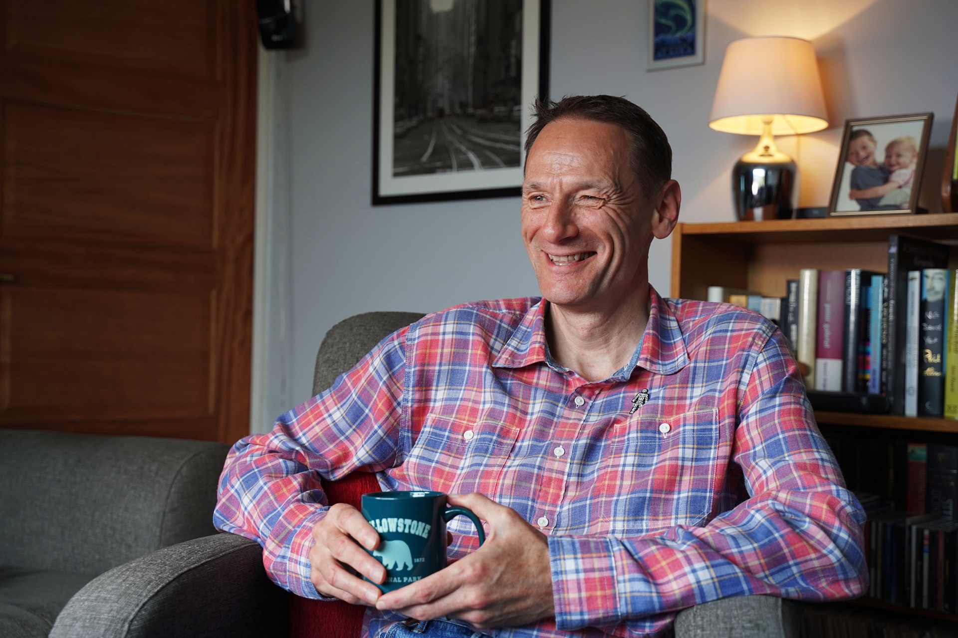 A smiling man wearing a red and blue shirt sitting in an armchair and holding a mug of tea.