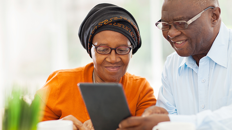 A woman wearing an orange top and a man wearing a white shirt look at a tablet device together