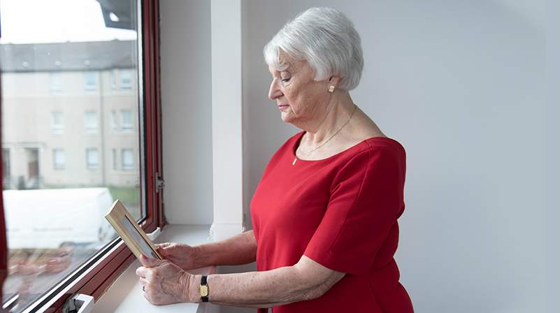 A woman standing by a window, looking at a photo frame