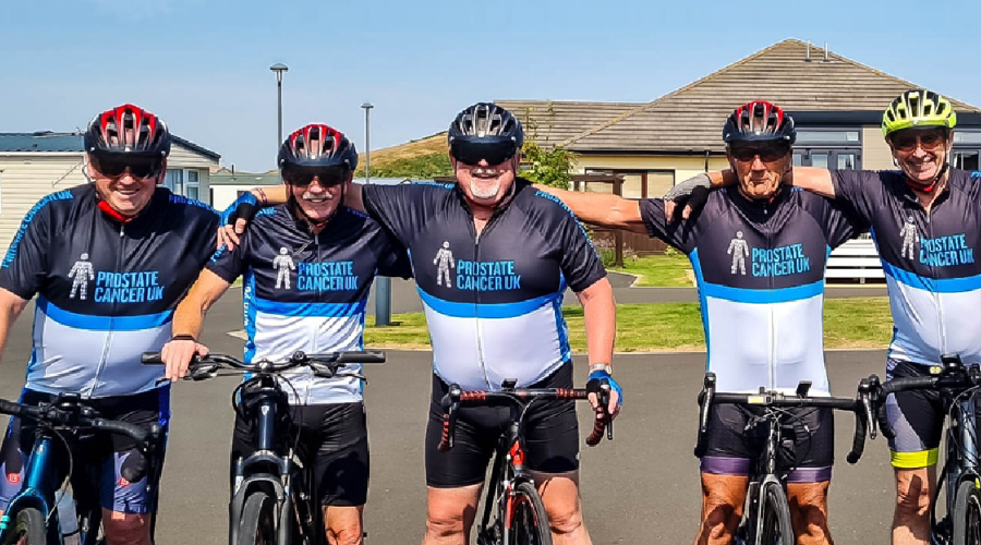 Five men standing arm in arm over their bikes, wearing blue and black Prostate Cancer UK branded cycling clothing
