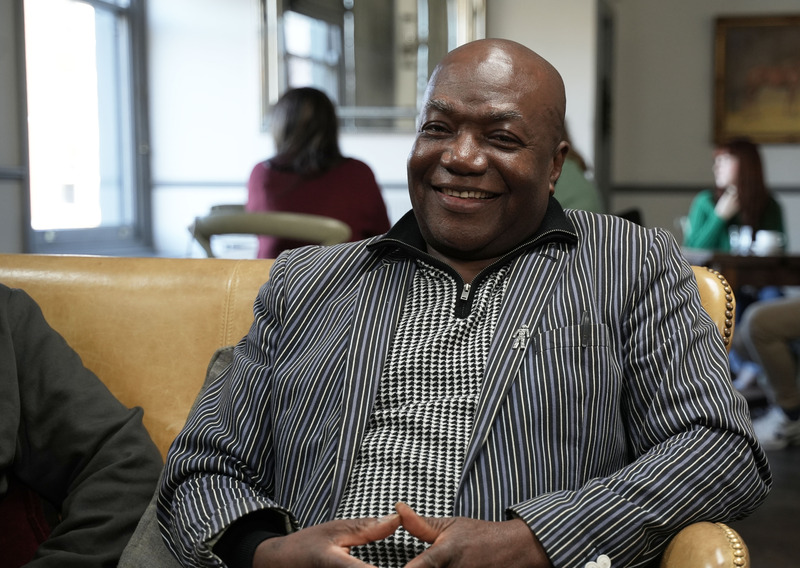 A man wearing a dark jacket, sitting on a sofa and smiling.