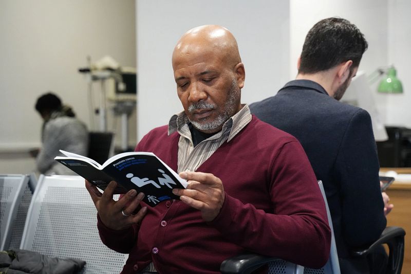 A man wearing a red cardigan sitting in a GP surgery waiting room reading a prostate cancer information pamphlet.
