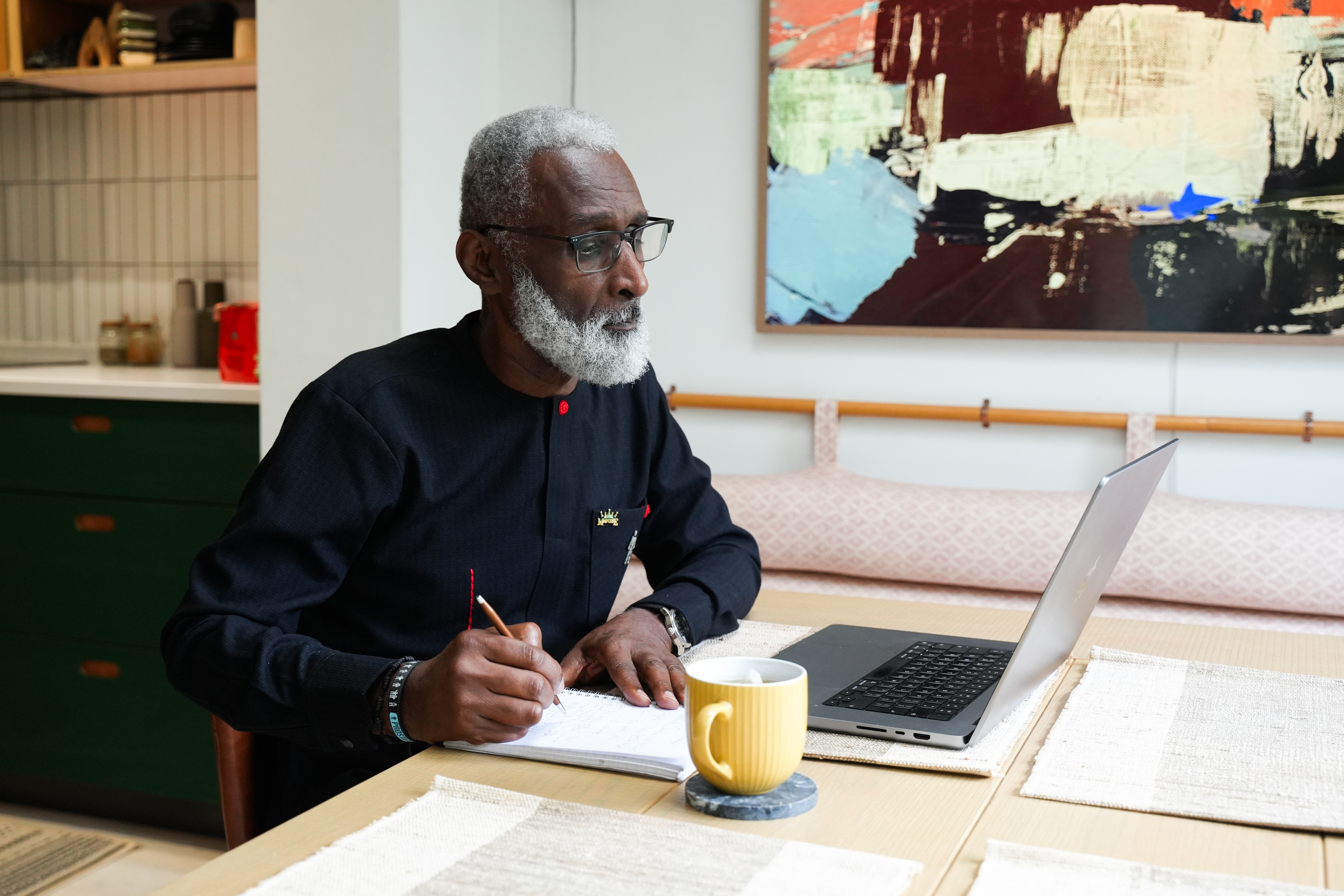 A man wearing a black shirt reading on a laptop and taking notes.