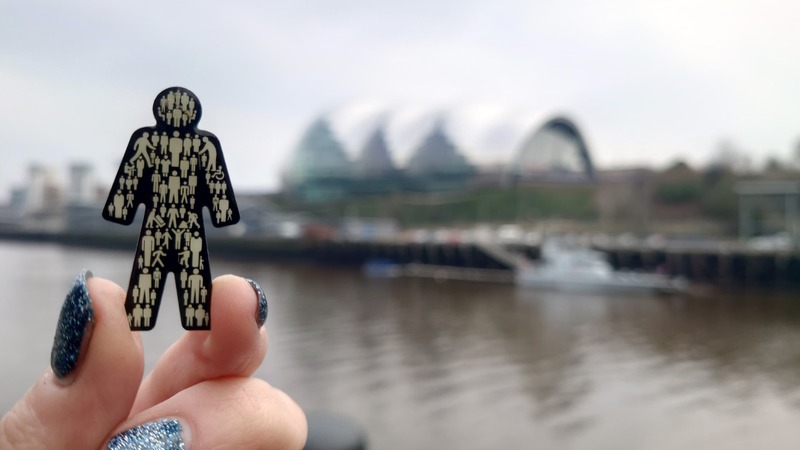 A close-up of a hand holding up a man-shaped pin bage, with Newcastle's Utilita Arena in the background