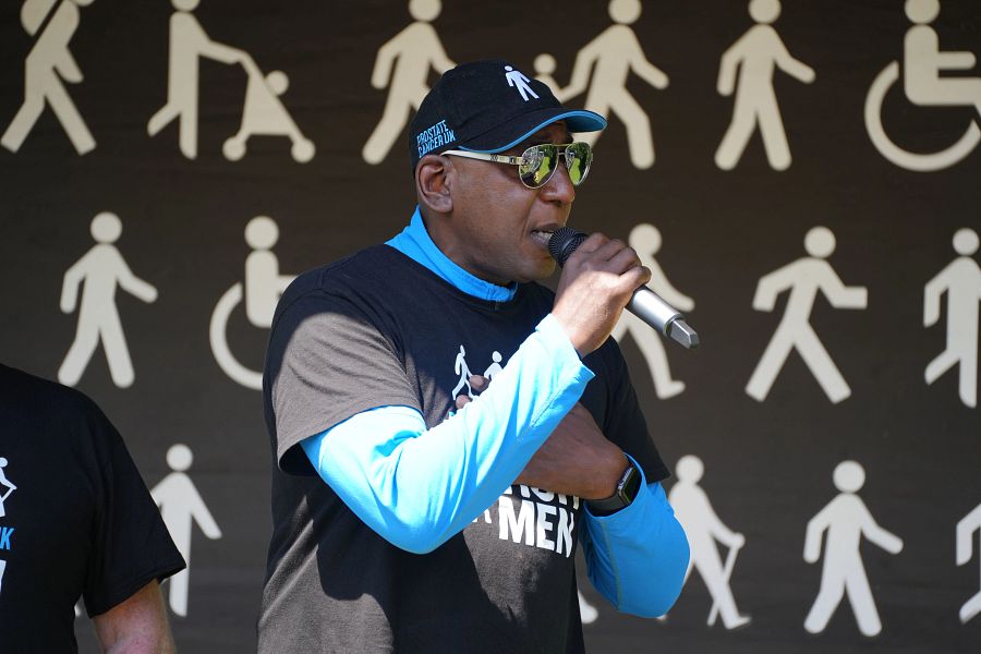 A man wearing a black Prostate Cancer UK t-shirt and cap holding a microphone and standing on a stage