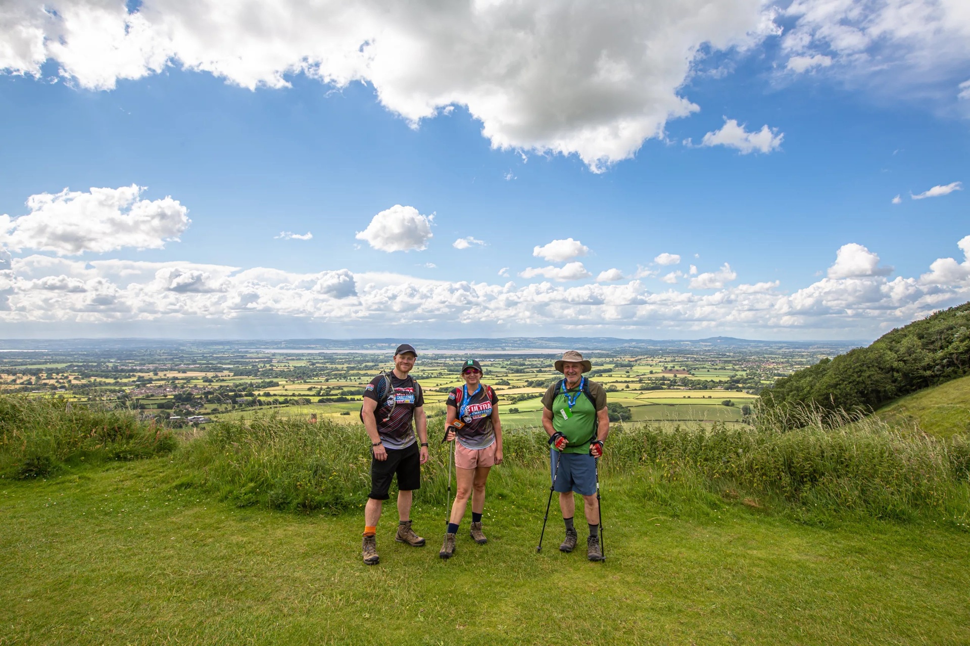 A woman and two men wearing walking gear standing on a grassy verge and smiling, with a hilly landscape in the background