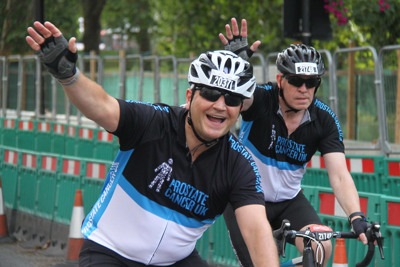 Two men wearing Prostate Cancer UK branded cycling kit wave and smile while riding by on their bikes