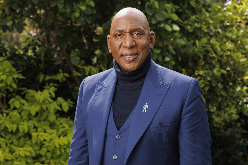 Actor Colin McFarlane smiles while wearing a blue blazer with Prostate Cancer UK's 'Man of Men' badge on the lapel.
