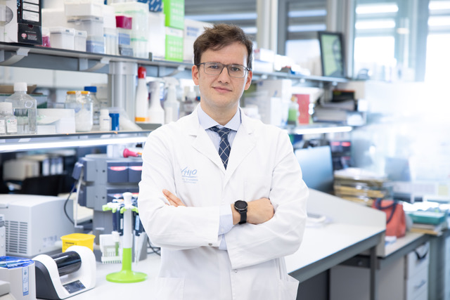 A man wearing glasses and a white lab coat standing in a science lab with his arms folded