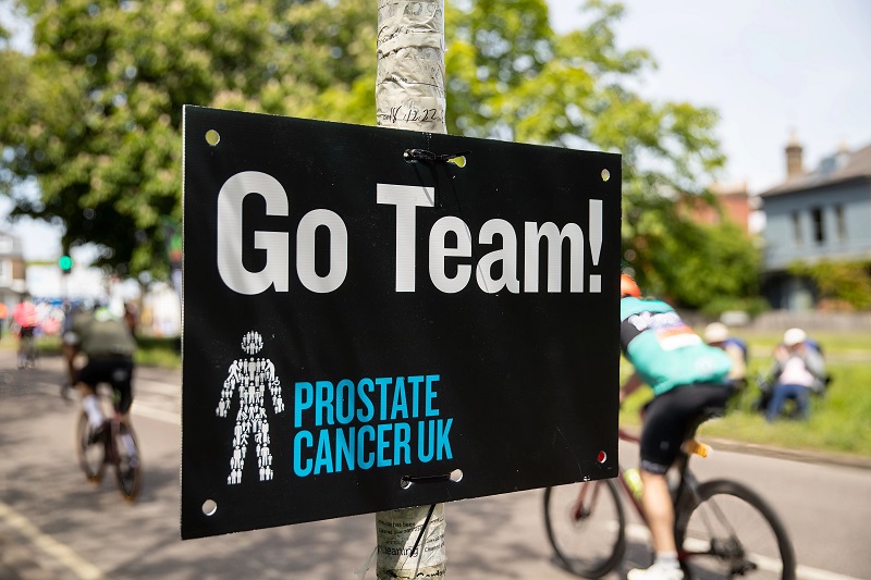 A sign reading 'Go Team!' with the Prostate Cancer UK logo, pinned to a tree, with cyclists riding along a rode in the background