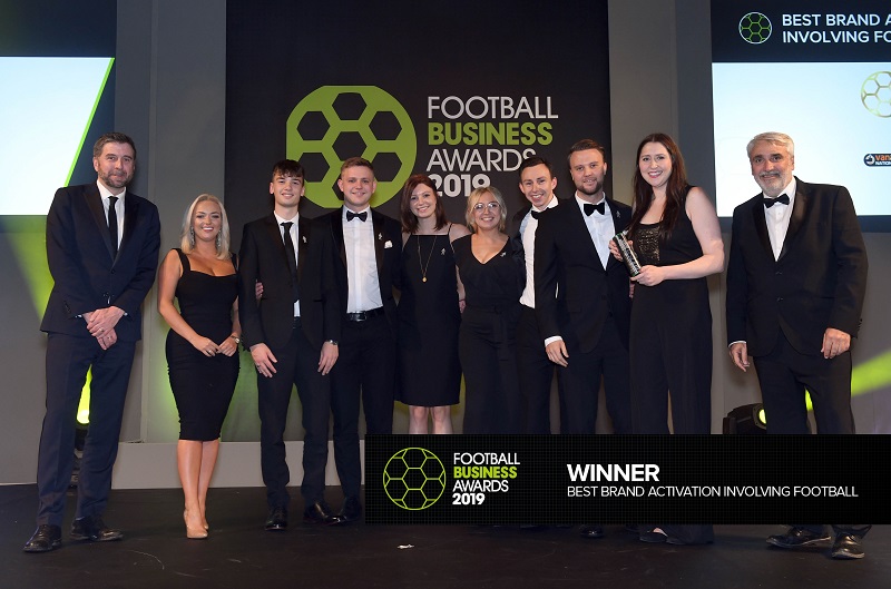 A group of 10 people in evening dress celebrate after receiving an award on stage.