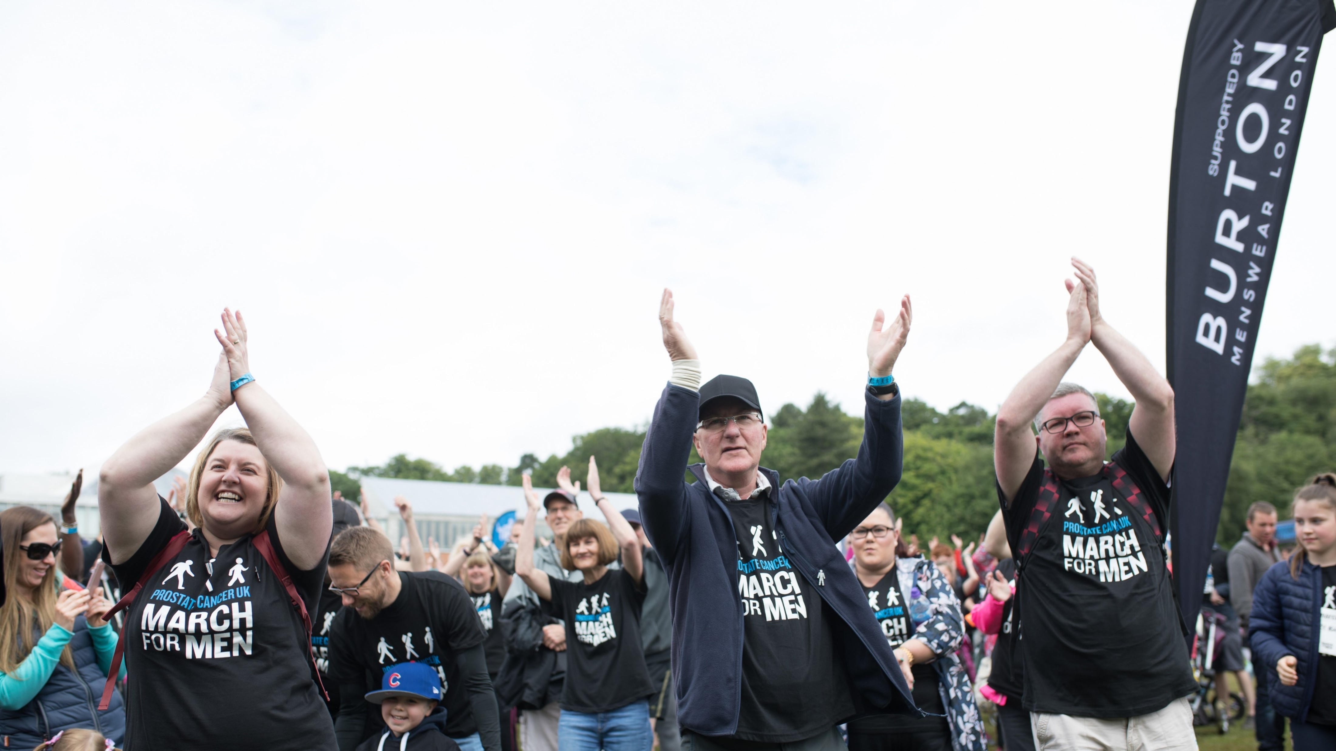 Two men and a women raise their hands and clap while warming up for a fundraising event in a park.