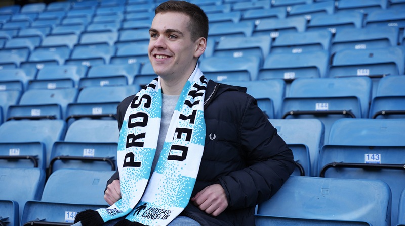 A young man sitting on a seat at a football ground wearing a Prostate Cancer UK scarf