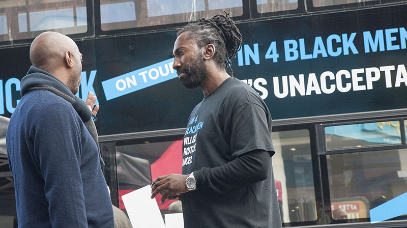 A man wearing a black t-shirt saying '1 in 4 Black men will get prostate cancer' talking to a member of the public. 