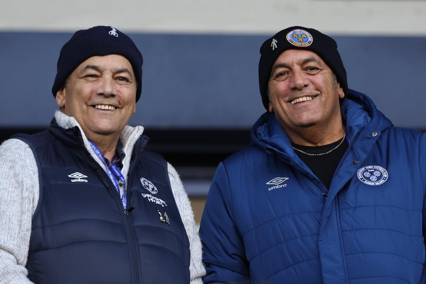 Two men at a football match wearing coats and woolly hats, smiling and laughing together.