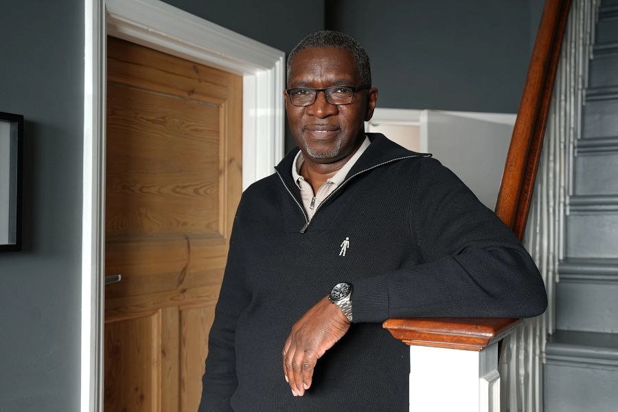A Black man wearing a blue sweater, standing in a hallway with his arm resting on the bottom of a staircase
