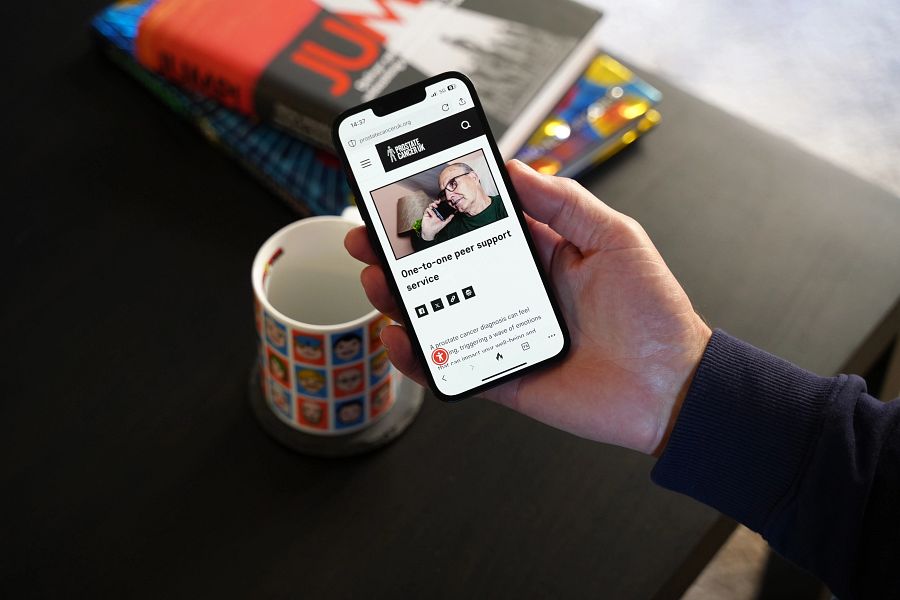 An outstretched hand holding a mobile phone showing the Prostate Cancer UK One-to-One Peer Support Service. 
