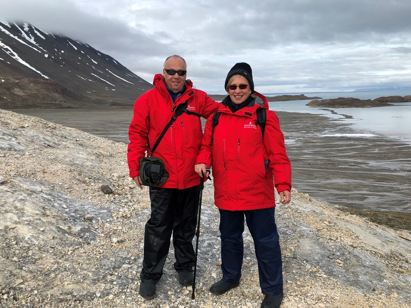 A man and a woman wearing red winter coats hiking in front of a mountainous terrain. 