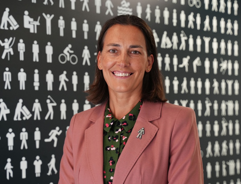 Chiara De Biase wearing a pink jacket and green shirt standing in front of the Prostate Cancer UK backdrop