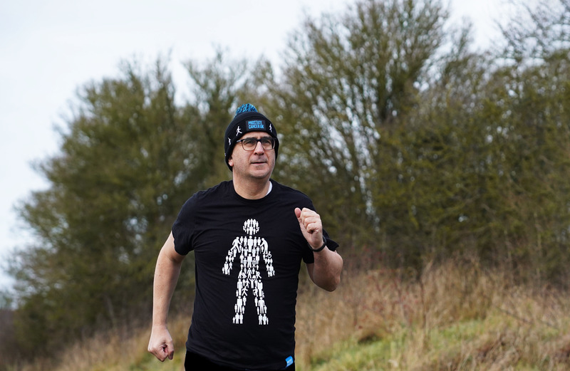 A man wearing a black Prostate Cancer UK t-shirt and a woolly hat out running in the countryside.