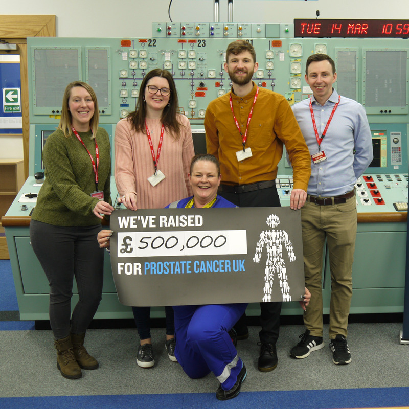 Three women and two men stand together and smile while holding up a giant cheque for £500,000.